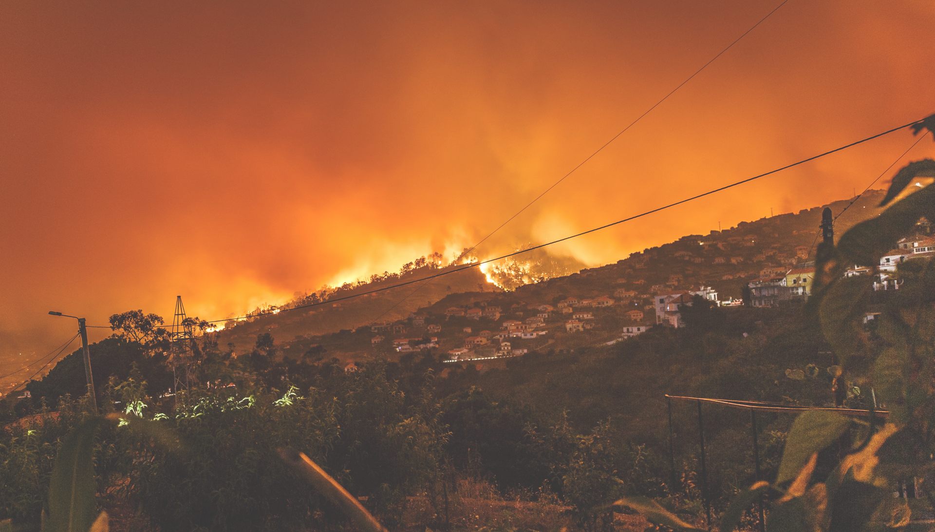 A wildfire advancing at night over a hillside village, the sky glowing orange